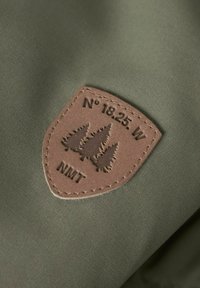 Maslinasto zelena tkanina s ušivenim smešnim kožnim pečatom koji prikazuje tri siluete drveta i tekst "N° 18.25. W NMT." Meka tekstura, oblik štita.