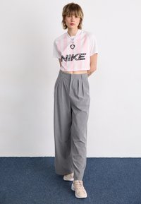 Rövidített fehér és rózsaszín csíkozott Nike póló, fekete logóval, szürke csíkozott, bőszárú nadrággal és bézs tornacipővel párosítva.