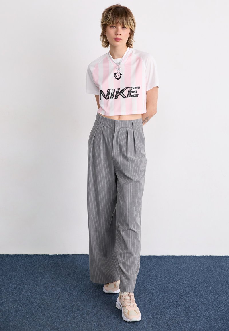 T-shirt Nike blanc et rose rayé coupé avec logo noir, associé à un pantalon large rayé gris et des baskets beiges.