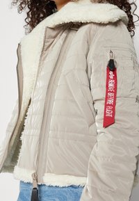 Beżowa ocieplana kurtka z kołnierzem ze sztucznego futra, zamkiem błyskawicznym i czerwonym uchwytem do suwaka. Na zewnątrz pikowany, teksturowany wzór, gładka podszewka.