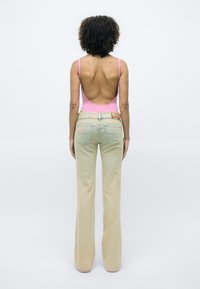 Femme aux cheveux bouclés portant un top rose dos nu et un jean beige à jambes larges, debout dos à un fond blanc uni.