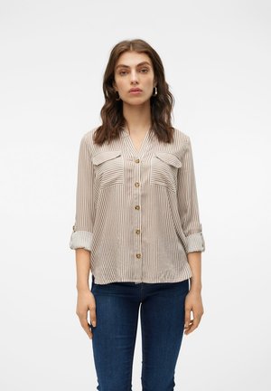 Femme aux cheveux bruns ondulés portant une chemise beige et blanche à rayures verticales avec boutons et manches retroussées ainsi qu'un jean bleu foncé, debout sur fond blanc.