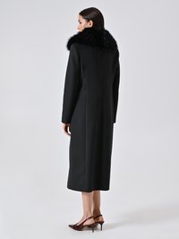 Vrouw in een lange zwarte jas met een grote zwarte bontkraag, in profiel staand met bordeauxrode slingback hakken op een effen achtergrond.