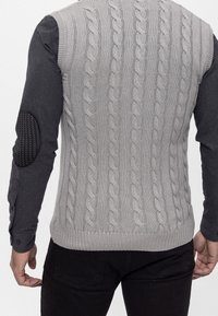 Grå strikket vest med kabelmønster, struktureret stof og ribkant forneden. Sorte albuebeskyttere på en mørk langærmet skjorte under.