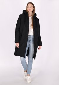 Sort hættetrøjeparka, knælang, med lynlås foran og sidelommer. Båret over en hvid skjorte og lyseblå jeans. Glat tekstur.