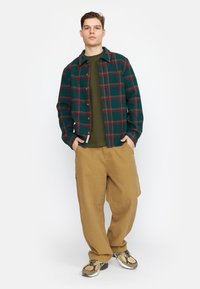 Geruite flanellen jas in groen en rood, lange mouwen, knoopsluiting, gecombineerd met een groene blouse, beige cargobroek en multicolore sneakers.