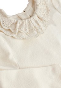 Vêtement couleur crème avec un col texturé à volants orné de dentelle délicate. Tissu doux avec une finition lisse.