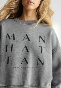 Grijze sweatshirt met vette zwarte letters waarin "MANHATTAN" en "NEW YORK CITY" staan, met een relaxte pasvorm en geribbelde halslijn.