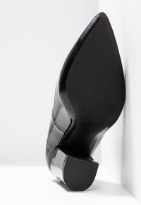 Chaussure habillée en cuir verni noir, à bout pointu et petit talon empilé, semelle tournée vers l'avant sur fond blanc.