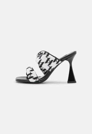 Mules con tacón en blanco y negro, con tiras de tela que presentan patrones en zigzag y un tacón puntiagudo negro. Textura suave con diseño de punta abierta.