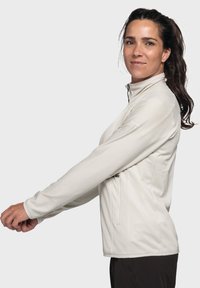 Leichte, crèmefarbene Zip-Jacke mit strukturierter Oberfläche, hohem Kragen und Seitentasche. Mit langen Ärmeln und taillierter Passform.