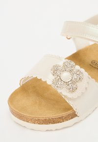 Witte leren sandaal met gegolfde banden en een glinsterende bloemaccent. Kurk voetbed met een beige zool.