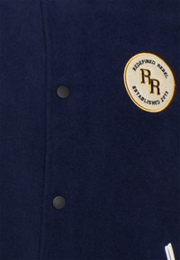 Marineblauwe wollen jasje met een ronde geborduurde patch met "REDESIGNED REBEL" en "ESTABLISHED 2013", aangevuld met zwarte knopen.