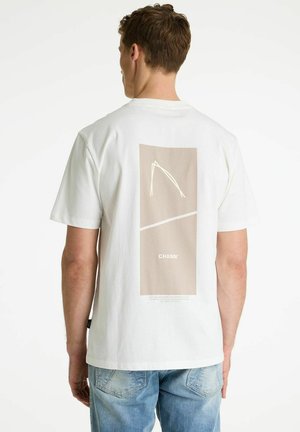 Mees kannab valget t-särki, millel on beež ristkülikukujuline graafika ja tekst "CHASIN'" seljal, koos helesiniste teksadega, vaatega seljale.