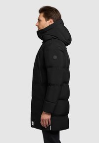 Zwarte gewatteerde jas met een hoge kraag en capuchon. Heeft een met rits afsluitbare borstzak, een zwart logo-opdruk op de schouder en een zijsplit.