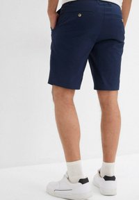 Pantaloni corti blu navy con taglio dritto, due tasche laterali e una tasca posteriore. Realizzati in un tessuto morbido e leggero. Indossati con scarpe da ginnastica bianche.