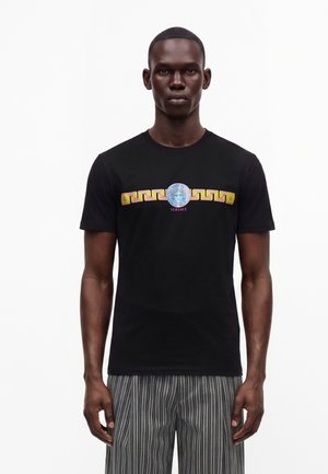 Uomo che indossa una t-shirt nera Versace con logo Medusa colorato e motivo geometrico sul petto, abbinata a pantaloni grigi a righe.