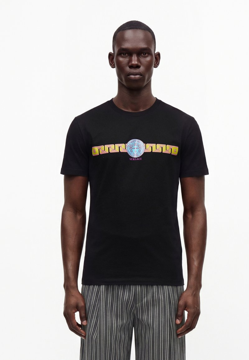 Άνδρας που φοράει μαύρο T-shirt Versace με πολύχρωμο λογότυπο Medusa και γεωμετρικό μοτίβο στο στήθος, σε συνδυασμό με γκρι ριγέ παντελόνι.
