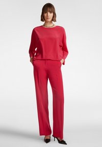 Blusa di raso rossa con scollo rotondo, abbinata a pantaloni larghi coordinati. L'outfit presenta tasche laterali ed è completato da tacchi neri.