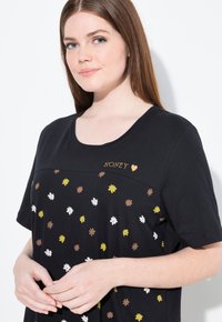 Camiseta negra con escote redondeado, estampado floral disperso en blanco, amarillo y marrón, con "HONEY" bordado en oro en el pecho.