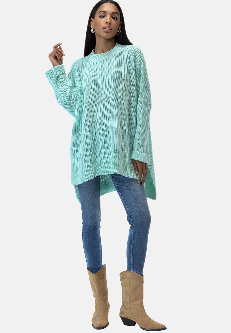 Hellblau gestrickter Pullover mit lockerer Passform, geripptem Ausschnitt, langen Ärmeln und asymmetrischem Saum, kombiniert mit eng anliegenden blauen Jeans und beigen Stiefeln.
