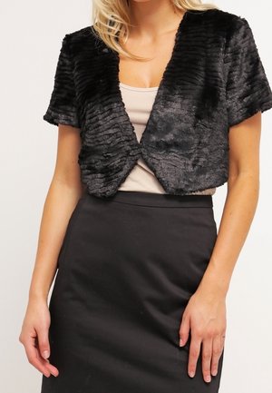Femme portant un boléro court en fausse fourrure noire par-dessus un haut beige et une jupe noire taille haute, debout les bras détendus.