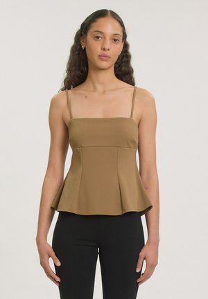 Vrouw met krullend haar die een bruine mouwloze peplum top en zwarte aansluitende broek draagt, staand tegen een effen achtergrond.