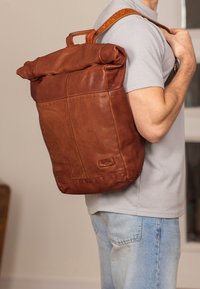 Brown leather backpack with a rolled top, featuring a textured finish and a side handle. Designed for comfort, with adjustable straps.