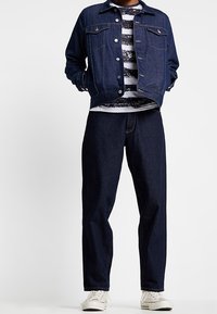 Džínová bunda v tmavě modré barvě se dvěma předními kapsami, kombinovaná s pruhovanou košilí s dlouhým rukávem a volnými tmavými džínami. Bílé tenisky celý vzhled doplňují.