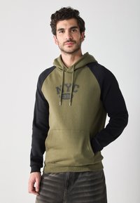 Sweatshirt à capuche avec un corps vert olive et des manches raglan noires. Il présente un graphique "NYC 1990", une poche avant et une capuche avec cordon.
