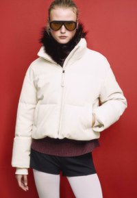 Weißer Daunenpark mit einem Kragen aus Pelzbesatz, Reißverschluss und seitlichen Taschen; kombiniert mit einem kastanienbraunen Pullover und schwarzen Shorts.