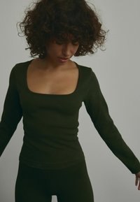 Haut long à manches longues en vert foncé, avec un décolleté carré, un design ajusté et un tissu texturé. Assorti avec un bas coordonné.