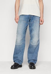 Blaue Jeans aus Denim mit lockerer Passform, versehen mit geschwungenen Nähten und Knie-Patche. Getragen mit schwarzen Schuhen und einem hellen T-Shirt.