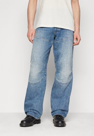Blaue Jeans aus Denim mit lockerer Passform, versehen mit geschwungenen Nähten und Knie-Patche. Getragen mit schwarzen Schuhen und einem hellen T-Shirt.