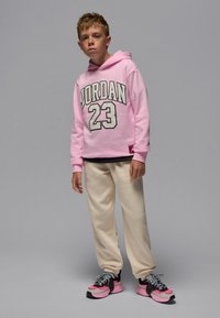 Rosa hettegenser med «JORDAN 23» i hvitt og grått, kremfargede joggebukser og svarte/rosa joggesko. Mot en grå bakgrunn.