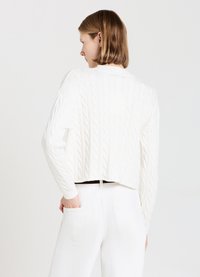 Weißer Kabelstrickpullover mit einem verkürzten Design, hohem Kragen und langen Ärmeln. Getragen über weißen, hochtaillierten Hosen.