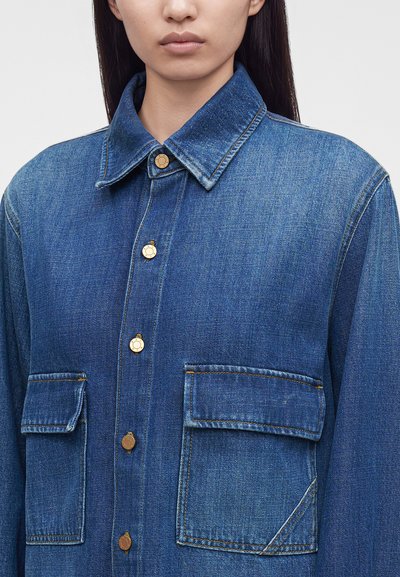 Femme portant une veste en jean bleue avec de grandes poches avant à rabat et des boutons dorés, montrée des épaules au menton sur un fond uni.