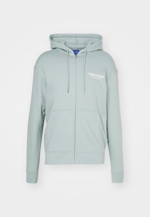 Homme portant un sweat à capuche zippé vert clair avec poches avant et jean gris clair, mains dans les poches, debout devant un fond uni.