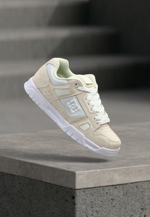 Baskets beige et blanches avec lacets et logo sur le côté, en suspension au-dessus d'un rebord en béton gris avec des marches floues en arrière-plan.