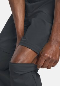 Handen die de ritssluiting van een zwarte broek naar beneden trekken om een bloot kniegewricht en het onderbeen van een donkerhuidig persoon te laten zien.