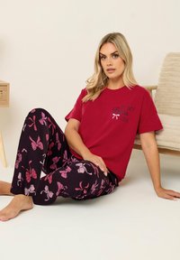 T-shirt en coton rouge avec le texte "DANS MA PÉRIODE Bow", associé à un pantalon noir avec des motifs floraux roses. Modèle assis sur un sol neutre.