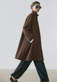 Woman walking wearing oversized brown buttoned coat, black pants, black slip-on shoes, and large black sunglasses against plain background.