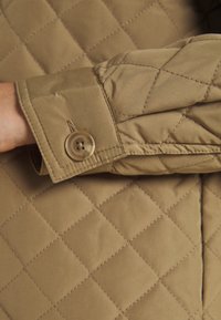 Een lichtbeige gewatteerde jas-sleeve met een knoopdetail. De textuur is glad, met ruitpatronen en een nette afwerking van de naden.