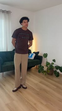 Mladý muž s kučeravými vlasmi stojí v obývacej izbe, má na sebe hnedé tričko a béžové nohavice, vedľa zeleného gauča a veľkej kvetináča s rastlinou.