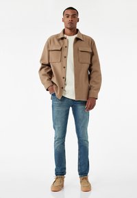 Chaqueta de lana color tostado con dos bolsillos frontales, llevada sobre una camiseta de color claro, combinada con jeans azules y zapatos de gamuza tostados.