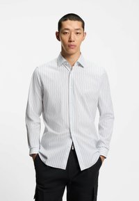 Hvid skjorte med knapper og lodrette blå striber, med spids krave og lange ærmer, kombineret med sorte bukser.