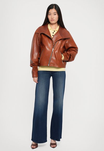 Chaqueta de motocicleta de cuero marr�ón con cuello ancho, cierre de cremallera y mangas oversized, combinada con una camisa amarilla de cuello y jeans de campana oscuros.