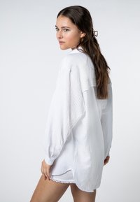 White oversized shirt in textured fabric, featuring long sleeves, a back yoke, and a hem with a small decorative trim.