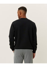 Felpa nera con scollatura rotonda e maniche lunghe, caratterizzata da una texture liscia. Indossata con pantaloni grigi chiari, vista da dietro.