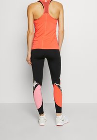 Korálový sportovní top s racerback z mesh detailů, kombinovaný s černými legínami s abstraktními růžovými a korálovými vzory a lehkými bílými teniskami.